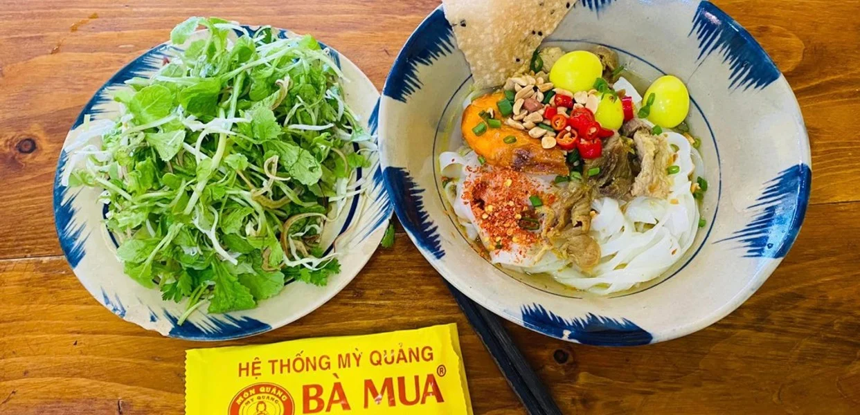 Top-down view of a bowl of Mi Quang Ba Mua in Da Nang, featuring yellow noodles, shrimp, quail eggs, peanuts, and a side of fresh herbs.
