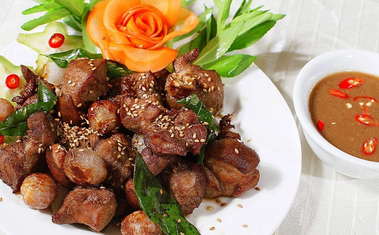 Close-up of a plate of Ninh Binh Mountain Goat meat (De Nui) garnished with sesame seeds and herbs, served alongside a bowl of traditional fermented soybean dipping sauce with chili