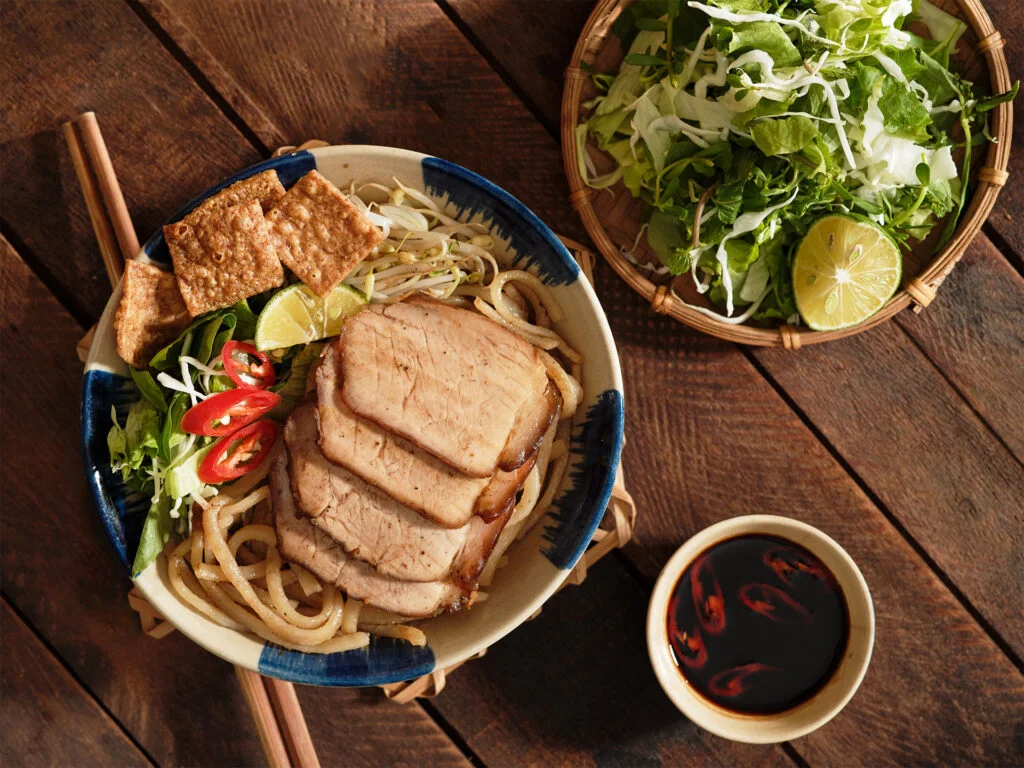 A bowl of traditional Cao Lau noodles with sliced pork, herbs, and crispy crackers.